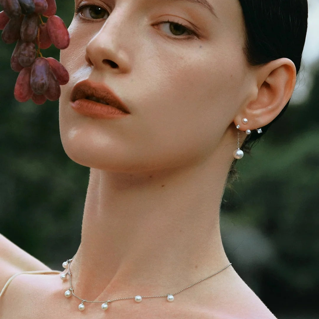A close-up lifestyle shot of a model wearing a delicate silver necklace and matching stud earrings with small white pearls, set against a soft, blurred green background.