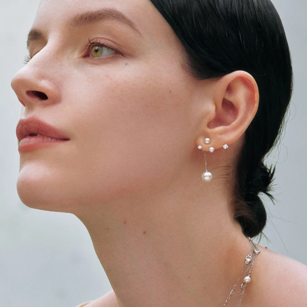 A close-up of a woman wearing a delicate silver-toned earring with small pearl-like beads and a dangling pearl drop, paired with a matching pearl necklace.