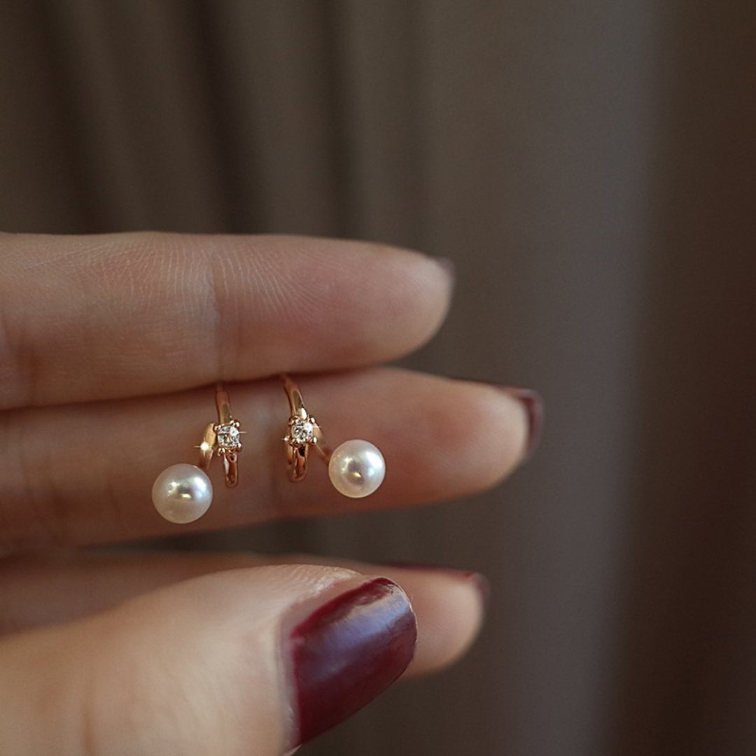 Two pearl diamond huggie earrings held between fingers against a neutral background