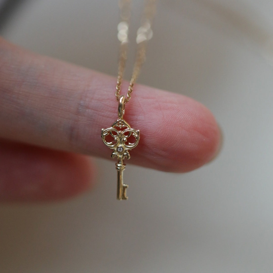Gold key-shaped pendant on a chain held between fingers against a neutral background
