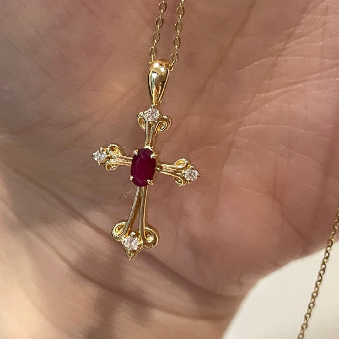Gold cross pendant with a red gemstone and diamonds on a hand.