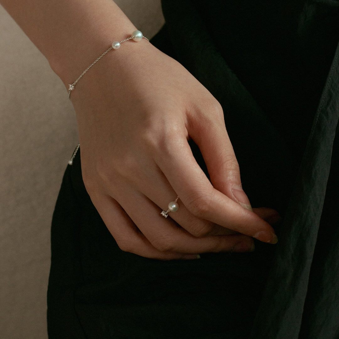 Hand wearing a pearl bracelet and chain ring on a dark background