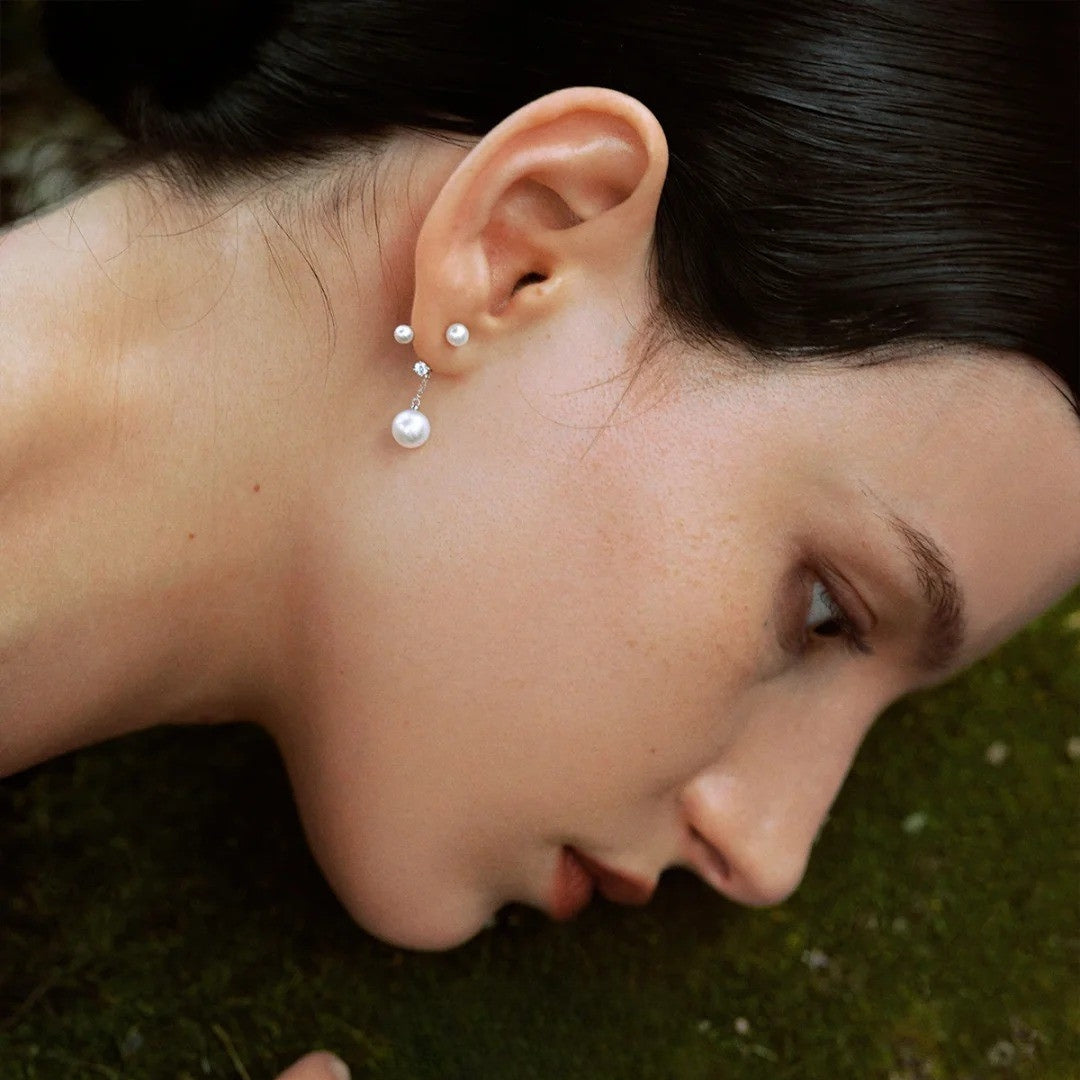 A close-up of a woman's ear wearing a pair of pearl stud earrings with small silver-toned posts and larger dangling pearls, set against dark hair and a green textured background.
