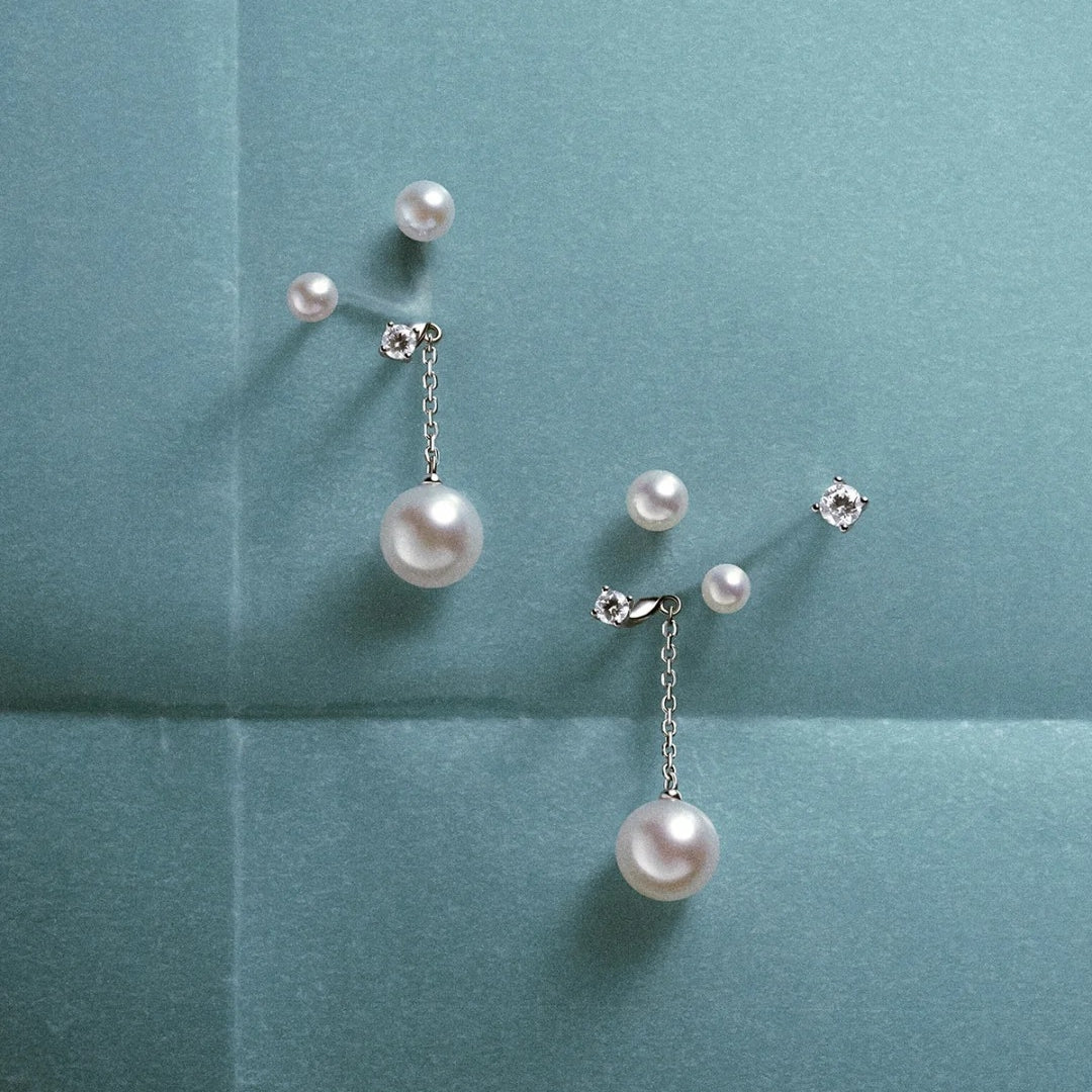 A pair of silver-tone drop earrings with large white pearls and small clear rhinestone studs, arranged on a textured teal background.