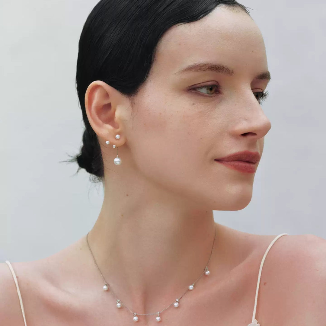 A close-up portrait of a woman with dark hair pulled back, wearing delicate pearl jewelry including a short pearl necklace, matching stud earrings, and a small drop earring against a soft light gray background.